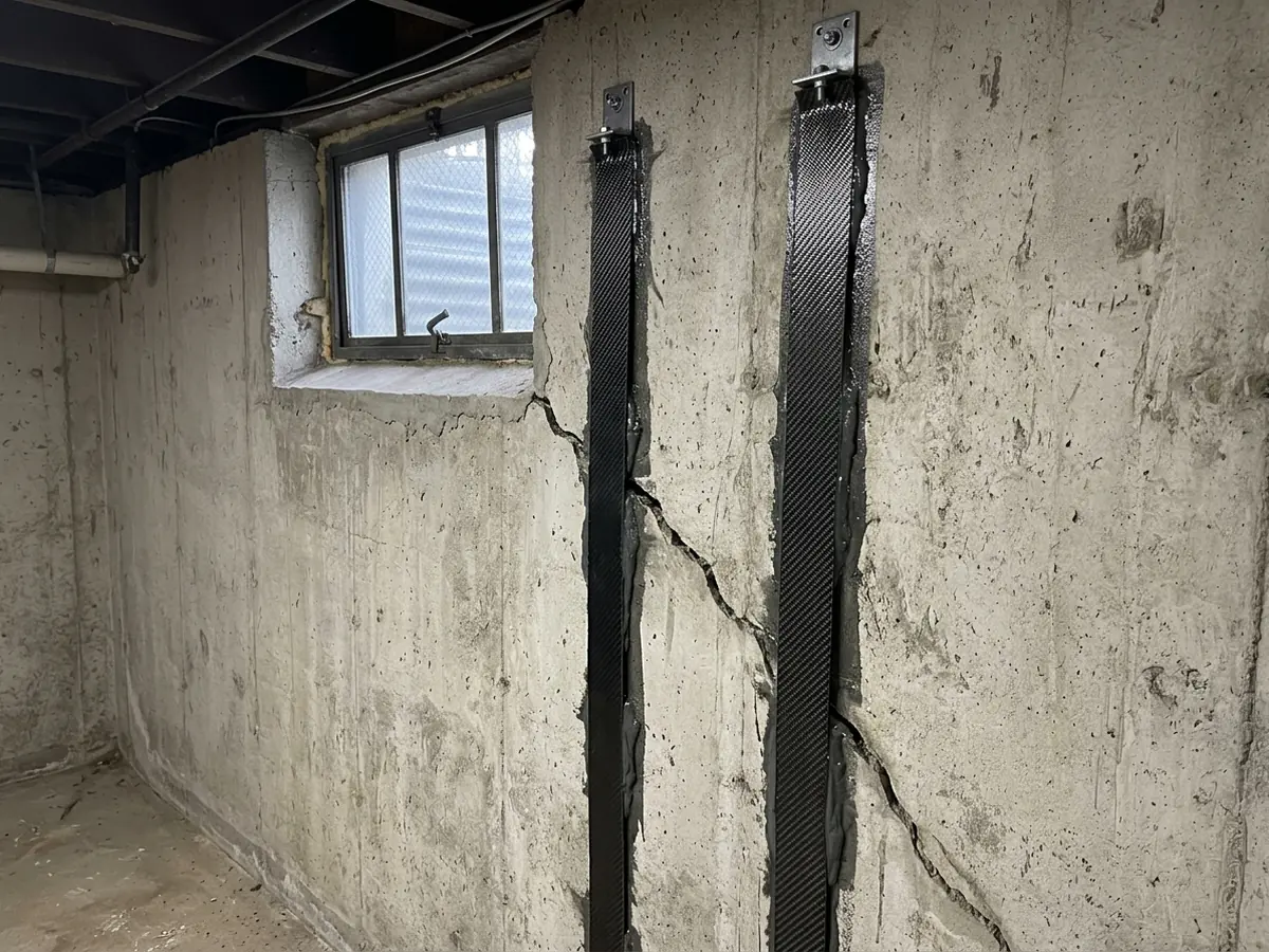Carbon fiber straps bonded across a diagonal crack on a poured concrete basement wall beside a small hopper window