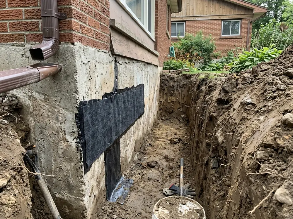 Narrow excavation beside a house foundation exposing a sealed crack in the poured wall under a peel-and-stick membrane