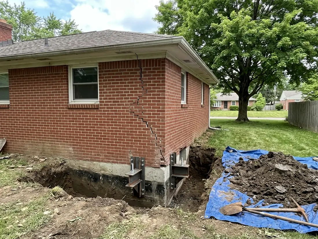 Dug-out corner of a brick ranch house with two helical pier brackets fastened to the footing, clay soil piled beside it